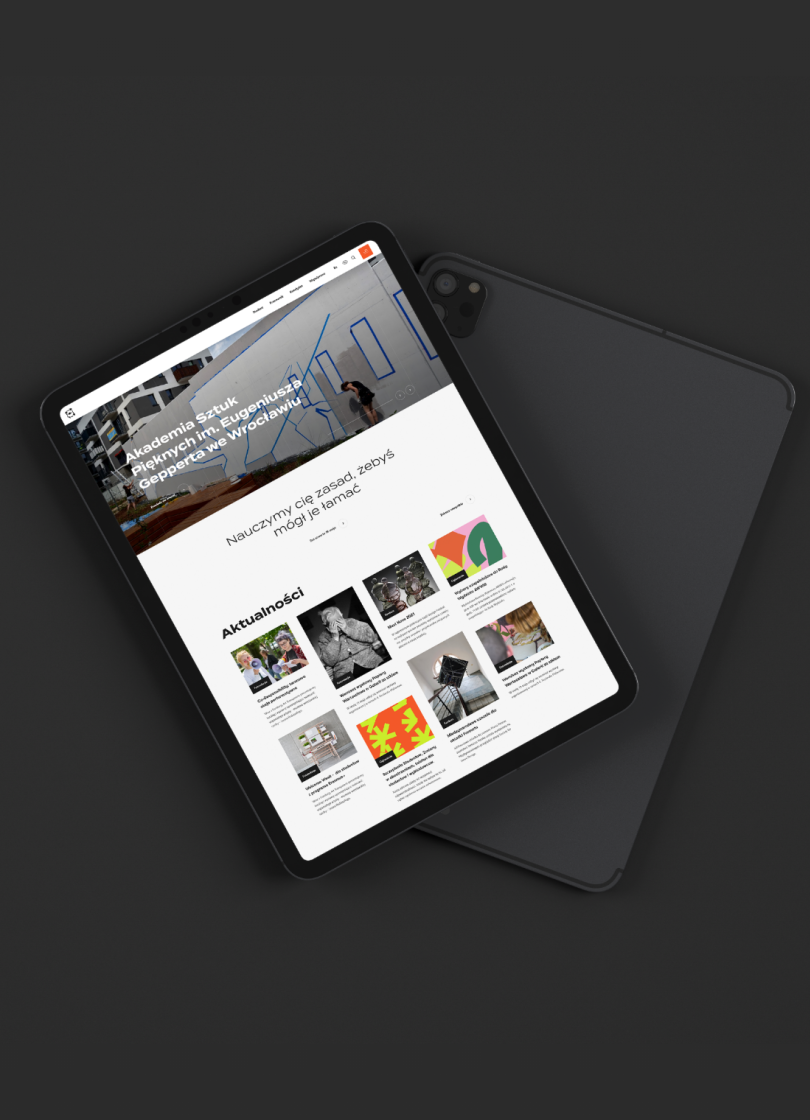 Tablet wyświetlający stronę internetową Akademii Sztuk Pięknych im. Eugeniusza Gepperta we Wrocławiu, z widocznym nagłówkiem, hasłem „Nauczymy cię zasad, żebyś mógł je łamać” oraz sekcją aktualności zawierającą miniatury artykułów i wydarzeń.