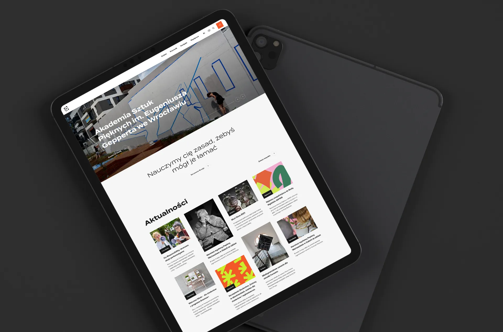 Tablet wyświetlający stronę internetową Akademii Sztuk Pięknych im. Eugeniusza Gepperta we Wrocławiu, z widocznym nagłówkiem, hasłem „Nauczymy cię zasad, żebyś mógł je łamać” oraz sekcją aktualności zawierającą miniatury artykułów i wydarzeń.